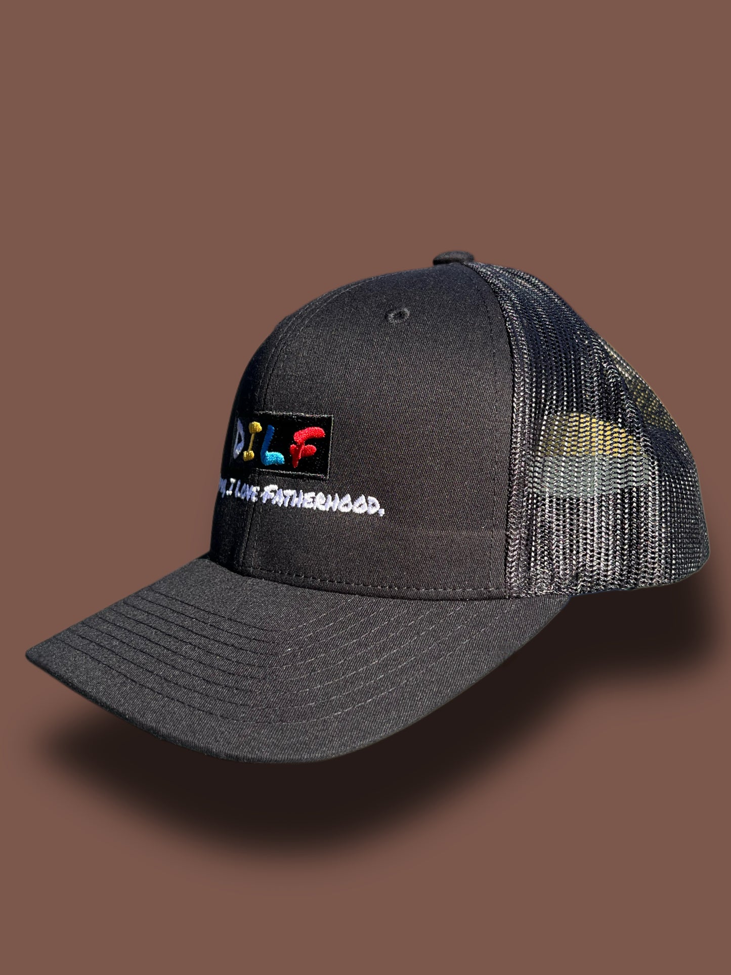 Side view of the classic DILF trucker hat with black front panel and embroidered D*mn I Love Fatherhood logo on a brown background