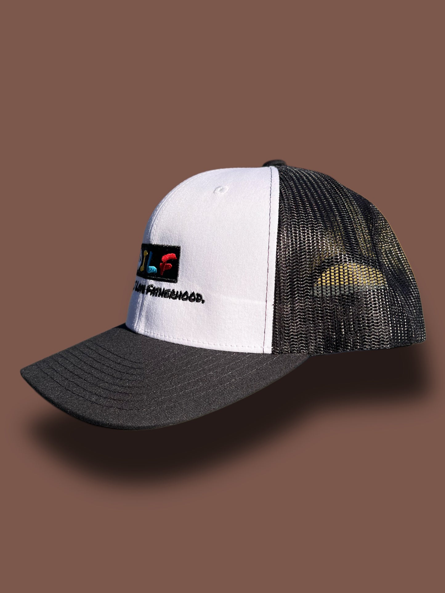 Side view of the classic DILF trucker hat with white front panel and embroidered D*mn I Love Fatherhood logo on a brown background